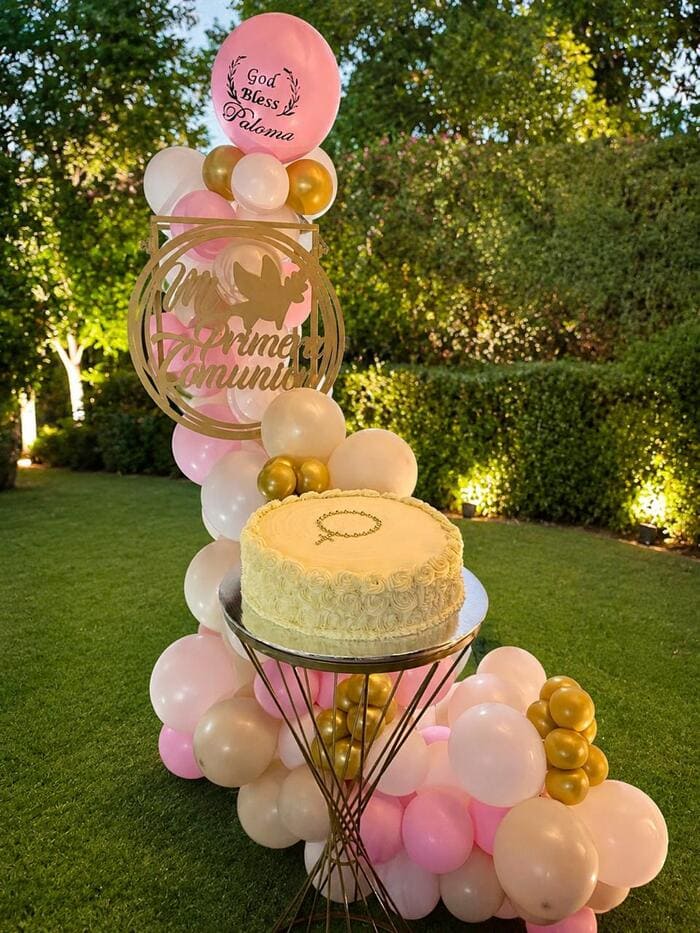 pastel de pimera comunion de Viry grc repostería sobre una mesa dorada y de fondo globos rosas en un jardín